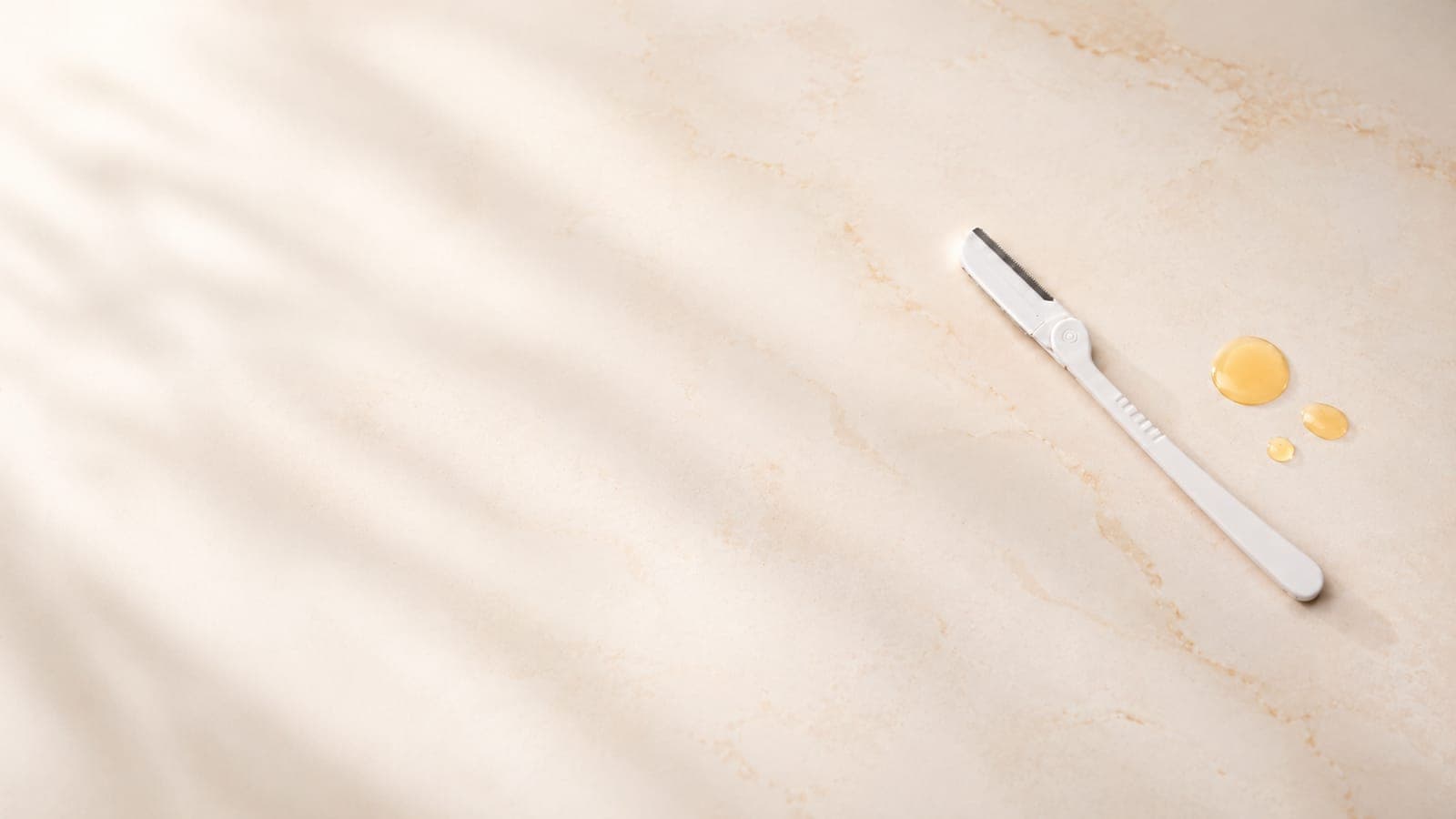 A jasclair dermaplaning razor beside drops of pre-shave oil on cream marble